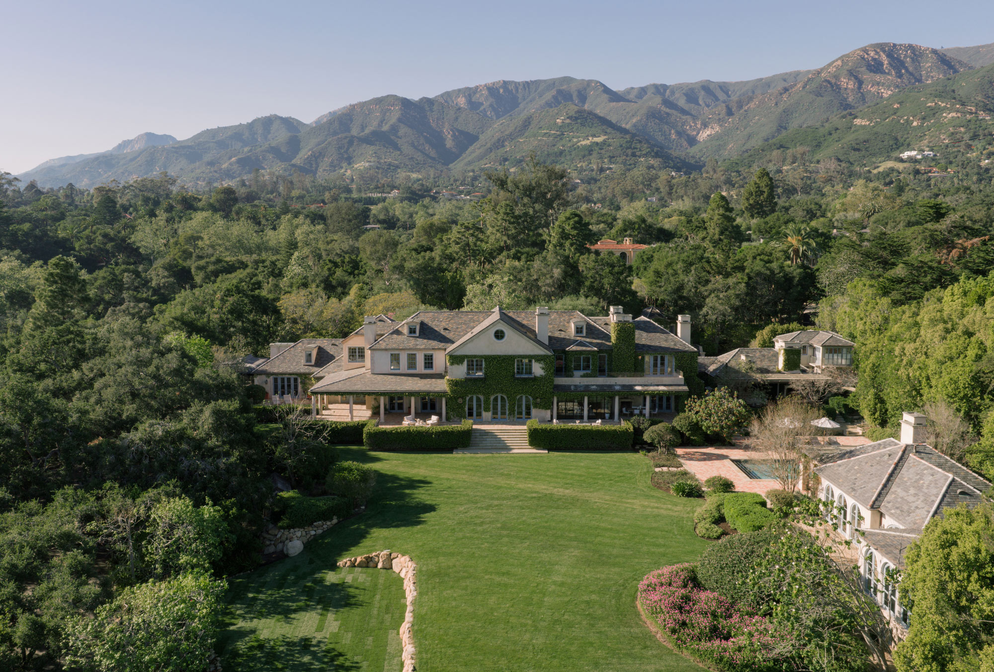 Aerial view of a sprawling luxury estate in Montecito with expansive green lawns, mature trees, elegant gardens, and the Santa Ynez Mountains rising in the background.
